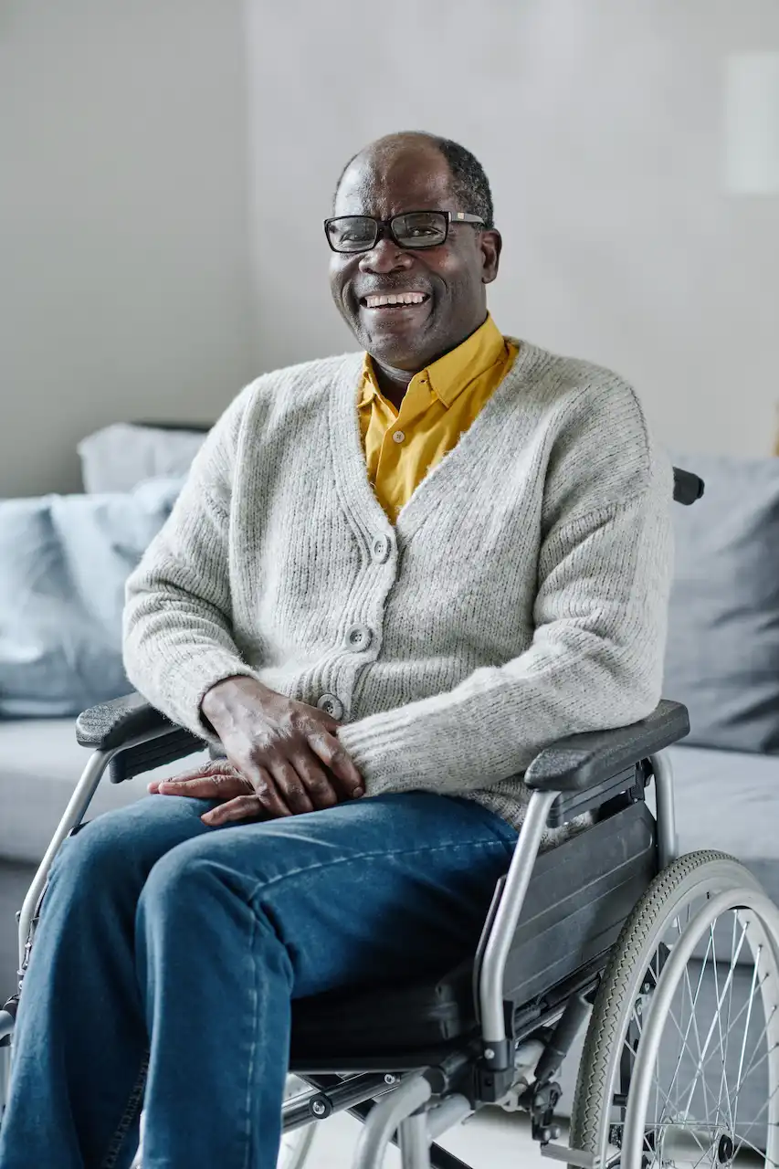 A mature man seated in a wheelchair, wearing a cardigan sweater and blue jeans.